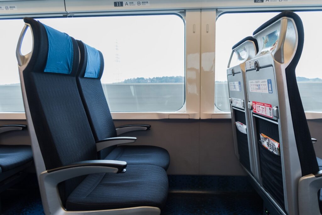 A pair of dark blue train seats with armrests and blue headrest covers inside a Keisei Skyliner train. The seats are positioned next to a large window, showing a blurred landscape of trees and hills in the background. The back of the seats in front features foldable tray tables and storage pockets containing informational pamphlets. The train's modern and clean interior is well-lit, with a sleek and minimalist design.