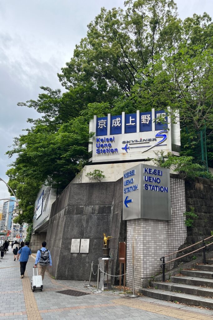 The entrance to Keisei Ueno Station in Tokyo is marked by a large blue and white sign with both Japanese and English text, including the Keisei Skyliner logo. The sign is mounted on a concrete wall with lush green trees behind it. Below, a smaller sign also points toward the station entrance. A golden statue of a bird stands on a pedestal near the stairs leading up to the station. Pedestrians, including a traveler with a suitcase, walk along the sidewalk under an overcast sky.