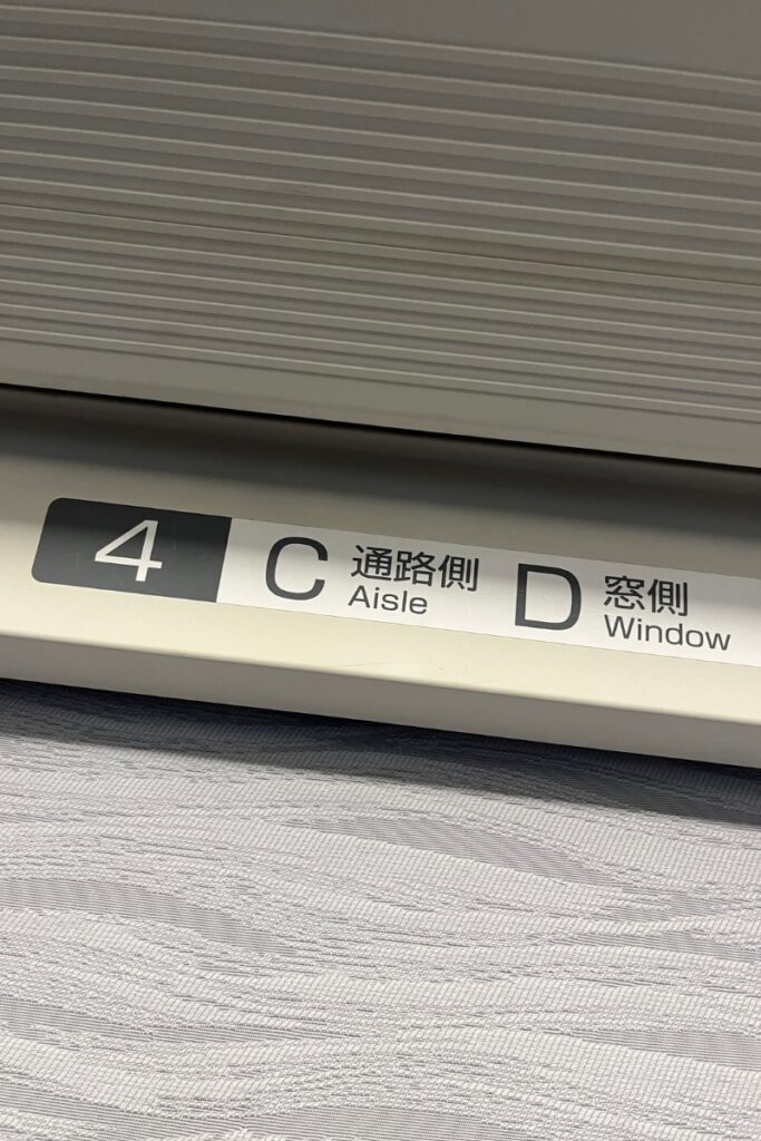 A seat number label inside the Keisei Skyliner, displaying "4C" for an aisle seat and "4D" for a window seat. The sign includes both Japanese and English text, with "通路側" (aisle side) and "窓側" (window side) indicating the seating arrangement. The label is affixed to a light-colored panel above the seats, with part of the textured seat fabric visible below.