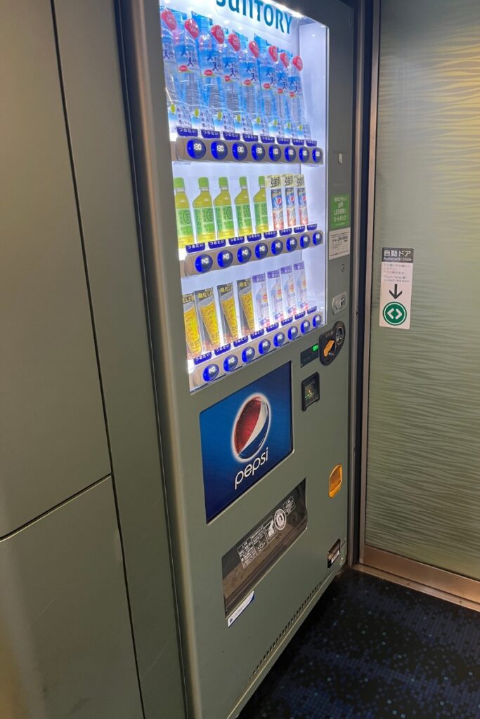 A vending machine inside the Keisei Skyliner train stocked with a selection of beverages, including bottled water, green tea, and canned drinks. The machine is branded with a Pepsi logo at the bottom and has a payment panel that accepts cash and electronic payments. Next to it, an automatic door with a sign indicating its function leads to another section of the train. The vending machine is illuminated, showcasing the available drink options with prices displayed below each row.