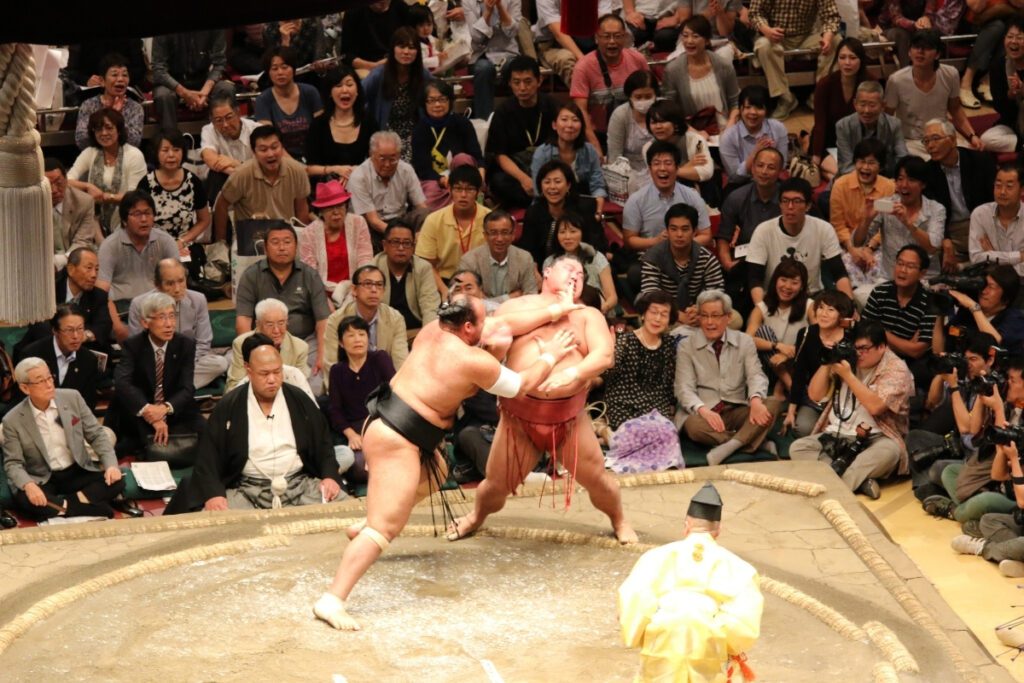 A sumo wrestling match in progress, with one wrestler in a black mawashi forcefully pushing his opponent in a red mawashi towards the edge of the ring. The wrestler in red leans backward, struggling to maintain his balance as the crowd watches with intense reactions. The audience includes a mix of photographers, fans, and formally dressed spectators, all closely following the bout. The referee, dressed in an elaborate yellow kimono, stands nearby, observing the fight. The expressions in the crowd range from excitement to awe, adding to the drama of the moment.