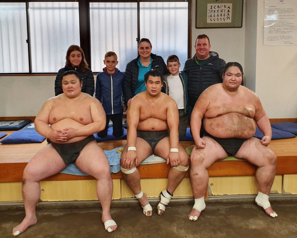 Our family poses for a photo with three sumo wrestlers after their training session in a sumo training facility. The sumo wrestlers, dressed in traditional mawashi, are seated on a wooden bench with bandaged feet, while the family, dressed in casual jackets and warm clothing, stands behind them, smiling. The setting includes blue training mats, a framed Japanese calligraphy piece on the wall, and a warm indoor atmosphere.