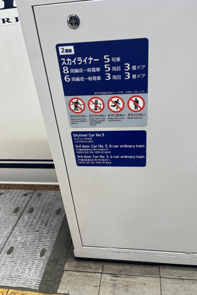 A platform sign at a Japanese train station displaying information for passengers about the Keisei Skyliner and ordinary train cars. The sign includes Japanese and English text, indicating the locations of Skyliner Car No. 5 and the third doors for ordinary trains with six or eight cars. Below the main text, four warning icons with accompanying messages advise passengers not to lean over the platform barrier, prop themselves against it, rush into the train, or enter restricted areas. The sign is mounted on a white panel near the platform edge, with tactile paving visible on the ground.