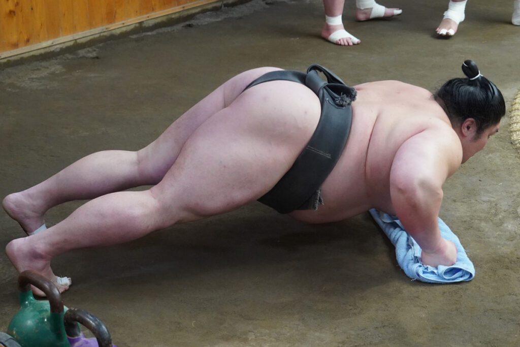 A sumo wrestler in a traditional black mawashi performs push-ups inside a sumo training stable. His hands are gripping a towel on the dirt floor, showcasing his strength and balance. Other sumo wrestlers' bandaged feet are visible in the background, along with kettlebells and wooden walls of the practice area.