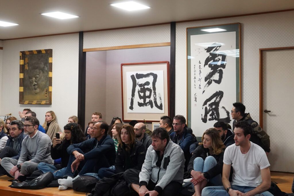 A group of spectators, including men and women of various ages, sits cross-legged on the wooden floor, attentively watching a sumo practice session. Some guests are taking photos, while others are focused on the training. The room features large framed calligraphy artwork on the walls and a bronze relief of a sumo wrestler, adding to the traditional atmosphere.
