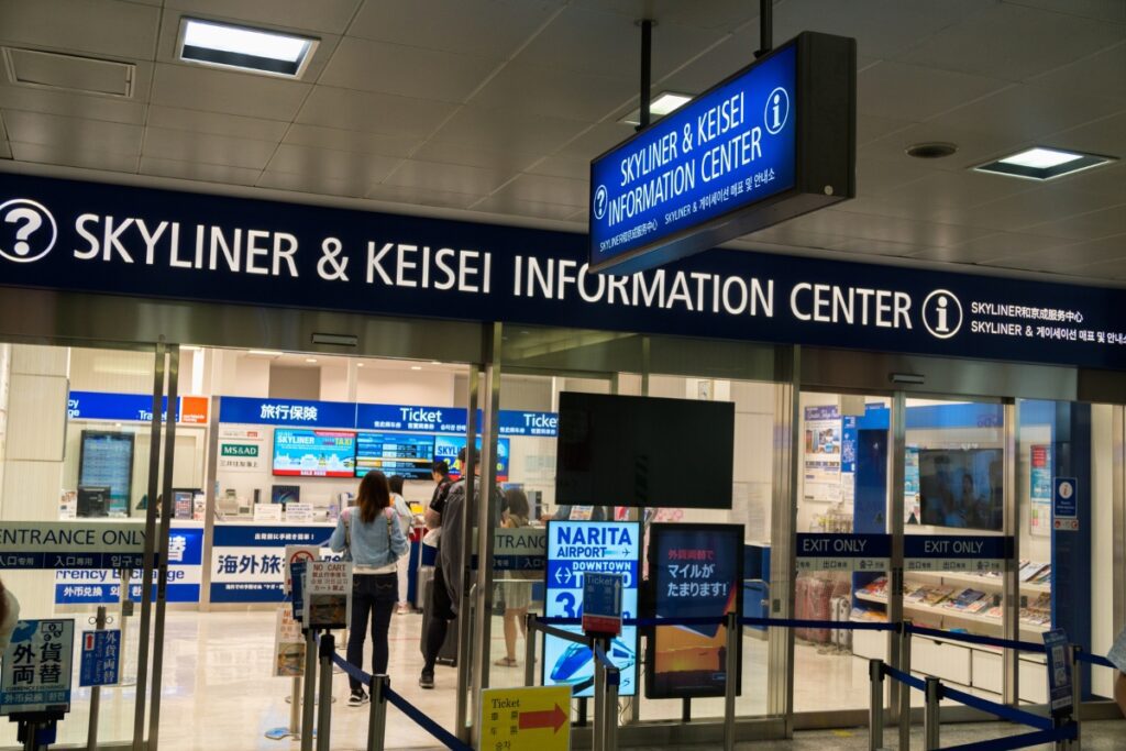 The Skyliner & Keisei Information Center at Narita Airport features a bright blue sign with white lettering above the entrance. The glass doors reveal a ticket counter inside, where several passengers are waiting in line to purchase tickets. Digital displays and signs inside provide travel information, including details about the Skyliner express train to downtown Tokyo. Additional signage near the entrance advertises a currency exchange service. The area is organized with barriers guiding customers towards the counter, and an "Exit Only" sign is visible on the right side of the entrance.
