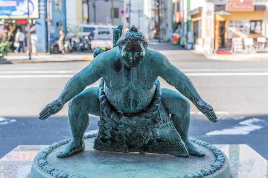 A bronze statue of a sumo wrestler in a low stance with arms extended is displayed on a round pedestal. This sculpture is located in Ryogoku, Tokyo—Japan’s historic sumo district—celebrating the city’s deep connection to the national sport.