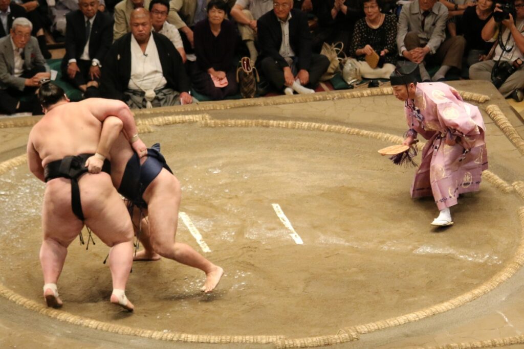 Two sumo wrestlers grapple inside the clay ring during a match, while the gyōji, the traditional sumo referee dressed in an ornate pink kimono, crouches nearby to closely observe the action. Spectators seated in the front rows intently watch the intense moment as the match unfolds.