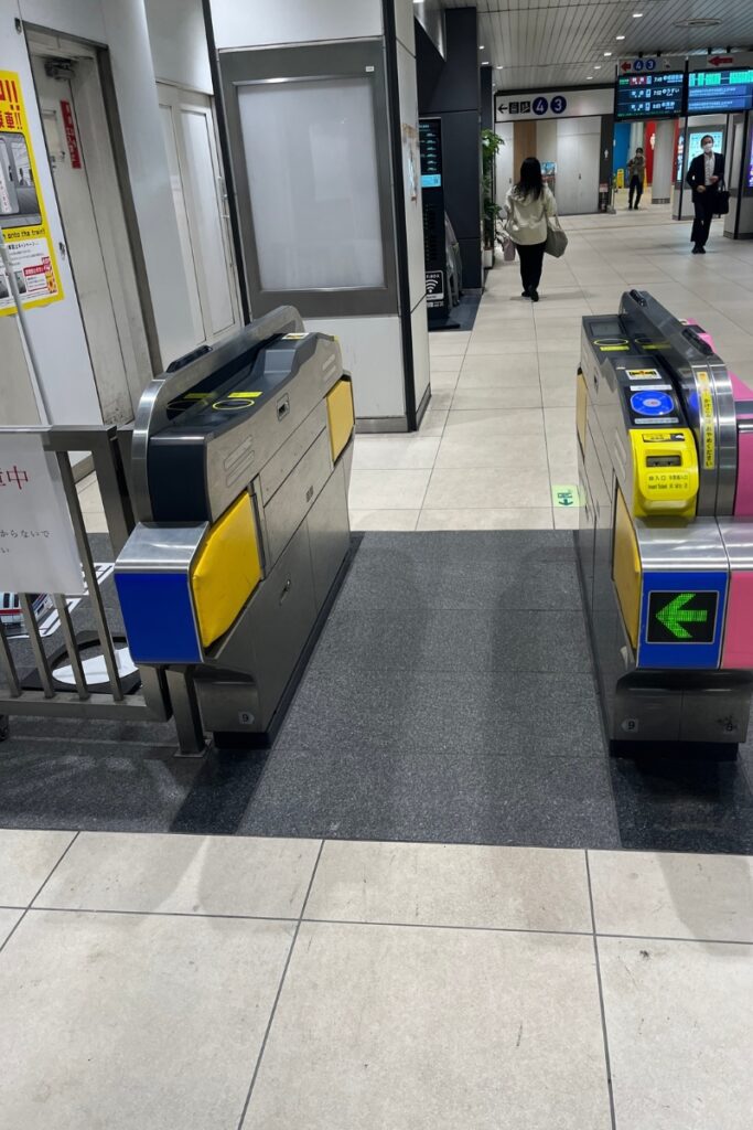 A set of automatic ticket gates at a Japanese train station, with a wide, accessible entryway in the center. The gates have touchpoints for IC cards and slots for paper tickets. A green arrow on the right gate indicates it is open for entry. In the background, passengers walk through the station, with directional signs above them displaying platform numbers and train information. The station interior is clean, with tiled floors and bright lighting.