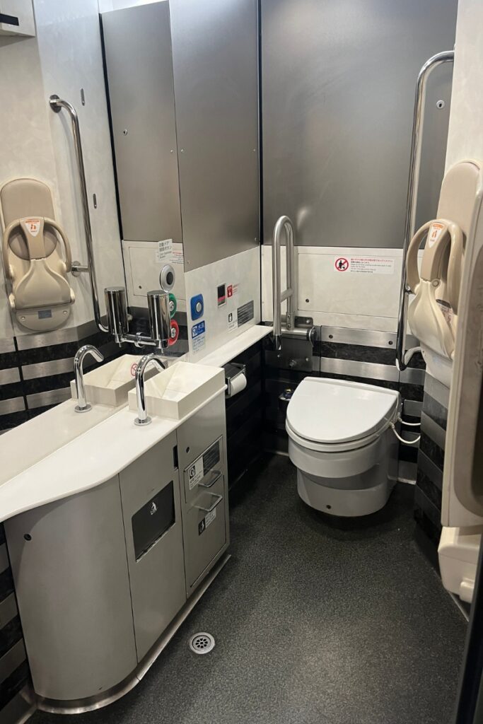 A spacious and accessible restroom inside the Keisei Skyliner train, featuring a modern toilet with safety grab bars, a dual sink with chrome faucets, and a baby seat mounted on the wall. The interior has stainless steel panels, black and white accents, and multiple accessibility features, including emergency buttons and handrails. The floor is textured for safety, and there are clear instructional signs on the walls.