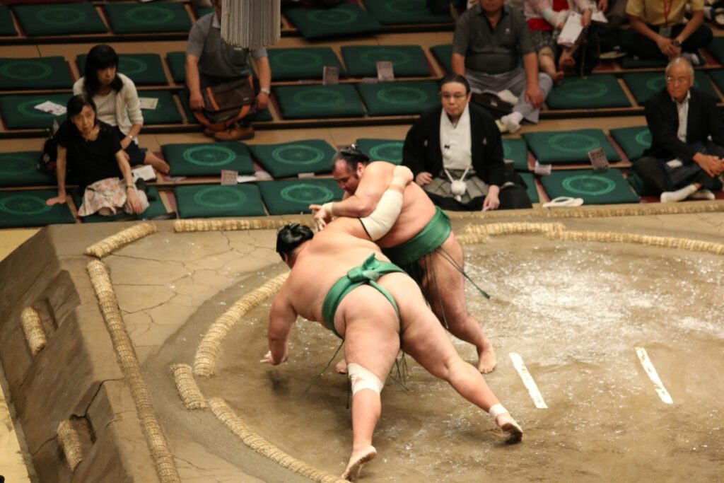 A sumo match captures the moment one wrestler, wearing a green mawashi, is being forced downward by his opponent, who is also clad in a green mawashi. The wrestler in control has his arm wrapped around his rival’s upper body, leveraging his weight to execute a tsukiotoshi (thrust down) technique. The wrestler being pushed down has both hands on the clay dohyo, bracing for impact. In the background, spectators seated on green cushions watch attentively, with some reacting to the action. A sumo elder, dressed in traditional black robes, observes from the side, maintaining a solemn expression. The dohyo’s straw bales mark the boundary of the ring, and the clay surface is sprinkled with traces of salt used for purification.