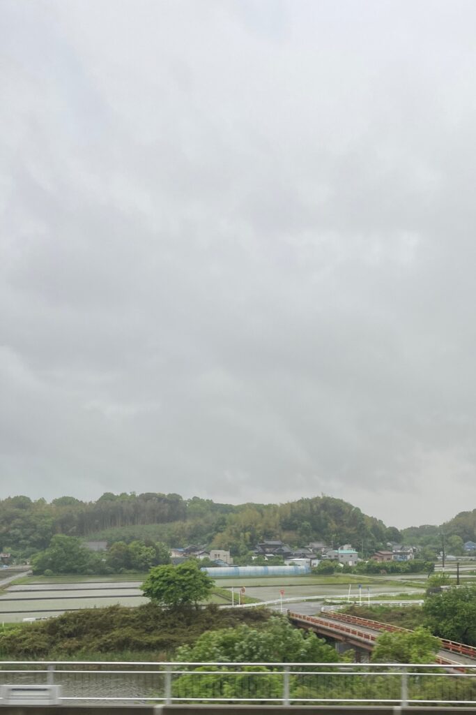 A scenic countryside landscape viewed from a moving train on a cloudy day. In the foreground, a metal guardrail runs parallel to the train tracks. Beyond it, lush green vegetation surrounds a small river, with a red bridge crossing over it. The middle ground features rice paddies reflecting the overcast sky, interspersed with rural houses and farm structures. In the background, rolling hills covered in trees and bamboo forests create a peaceful, natural setting. The sky is filled with thick, gray clouds, casting a soft, diffused light over the landscape.