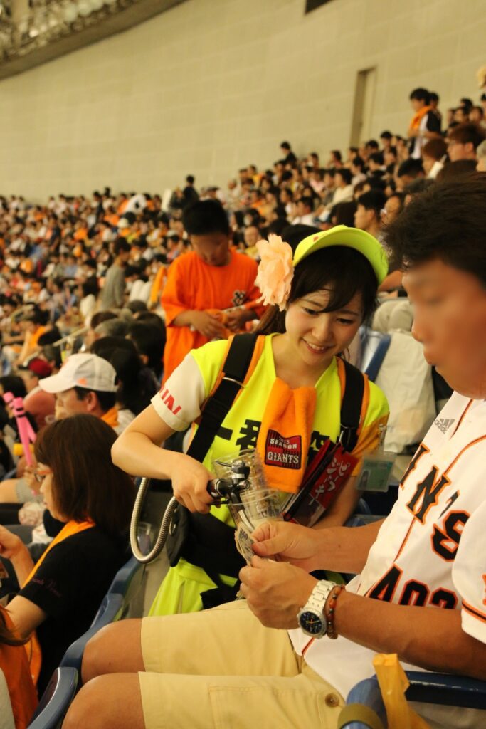 A smiling beer vendor in a bright yellow uniform serves a drink to a Yomiuri Giants fan in the stands during a game at Tokyo Dome, as a crowd of spectators fills the stadium seats in the background.