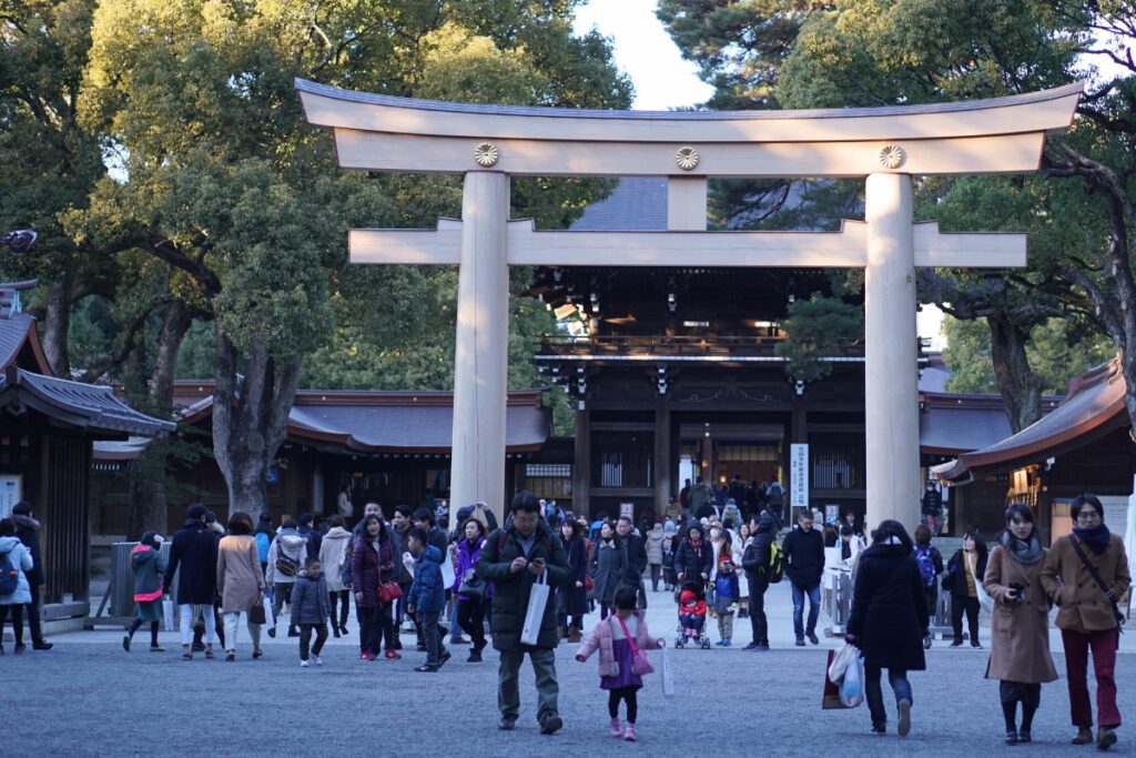 A large group of visitors walks beneath a grand wooden torii gate at Meiji Jingu Shrine in Tokyo, many dressed in coats for the cool weather as they make their way toward the main hall nestled among tall trees and traditional architecture.