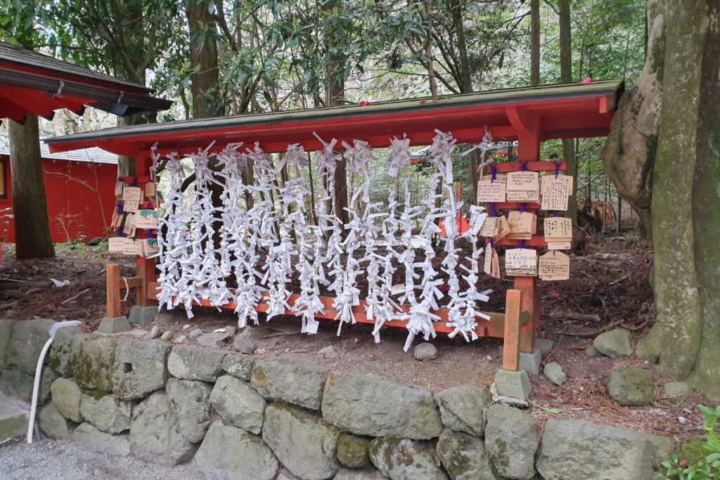 In a serene forest setting at Hakone Shrine, hundreds of omikuji fortune slips are tied to a red wooden rack, forming a curtain of paper representing hopes of leaving bad luck behind. On either side of the rack, wooden ema votive plaques hang with handwritten prayers and wishes left by shrine visitors. The tranquil surroundings and red shrine structures add to the spiritual atmosphere.