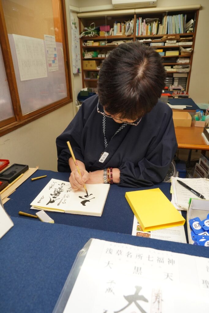 A person sits at a desk at Sensō-ji Temple in Asakusa, Tokyo, carefully writing traditional Japanese calligraphy in a goshuincho, a stamp book used for collecting temple and shrine seals. The room is lined with books, papers, and calligraphy tools, capturing the calm, respectful atmosphere of this cultural tradition.