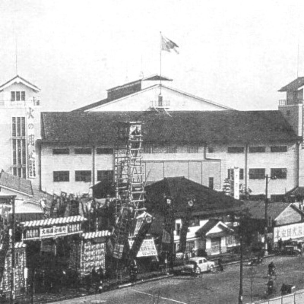 A black and white photo showing the exterior of the Kuramae Kokugikan sumo arena in Tokyo in 1950, with traditional banners, entrance decorations, and a Japanese flag flying above the roof, surrounded by early postwar street activity and vehicles.