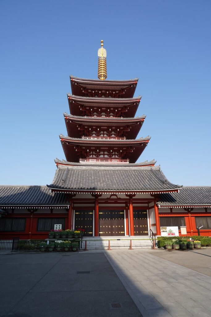 A striking five-story pagoda stands tall against a clear blue sky at Sensō-ji Temple in Asakusa, Tokyo, with its vibrant red wooden structure and ornate rooftop layers showcasing traditional Japanese Buddhist architecture.