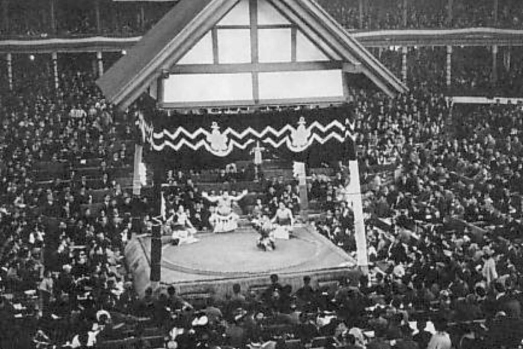 A black and white photograph showing the interior of the original Ryลgoku Kokugikan sumo arena in 1936. The venue is packed with spectators surrounding the central sumo ring, where a yokozuna performs a traditional dohyo-iri ring-entering ceremony under an ornate roof structure suspended above the ring.