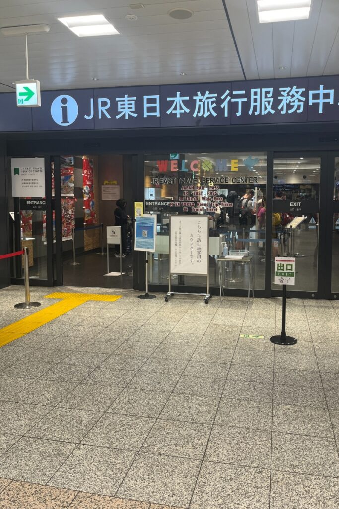 Entrance to the JR East Travel Service Center, with bilingual signage in Japanese and English. A line of travelers is visible inside, and various notices and signs are displayed at the front, including one promoting JR passes and limited express train tickets.