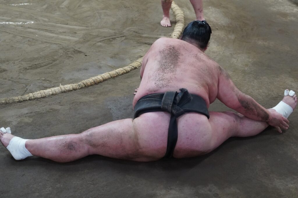 A sumo wrestler performs the “Matawari” stretching exercise inside the training ring, seated on the ground in a full straddle split while reaching toward his foot. His back and limbs are dusty from training, and his feet are wrapped in tape for support.