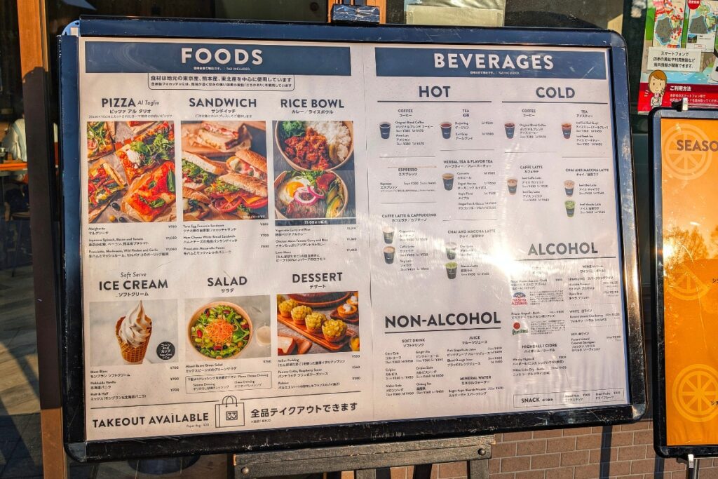 A bilingual café menu board from Café 33 displays a range of food and drink options in both English and Japanese, including pizza, sandwiches, rice bowls, salads, soft serve ice cream, desserts, and beverages such as coffee, tea, juice, and alcoholic drinks, with colorful images illustrating several food items.
