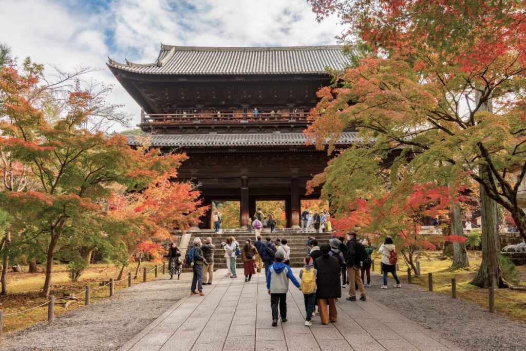 Visitors walk beneath vibrant autumn foliage toward the grand Sanmon gate at Nanzenji Temple in Kyoto, a towering wooden structure that serves as the iconic entrance to the historic Zen Buddhist complex.