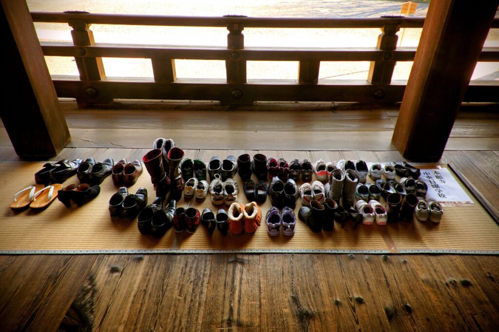 A neatly arranged collection of shoes sits on a tatami mat outside a wooden hall at Nishi Hongan-ji Temple in Kyoto, reflecting the cultural custom of removing footwear before entering sacred or traditional indoor spaces in Japan.