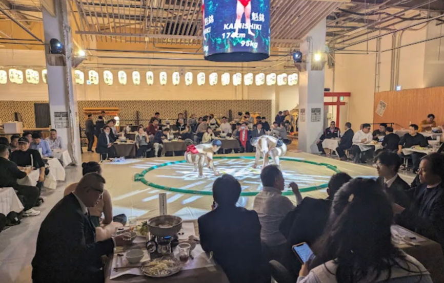 Guests dine around a central sumo ring inside a restaurant, watching two sumo wrestlers square off under a digital screen displaying stats for "Kanishiki" (172 cm, 95 kg). The space is warmly lit with Japanese lanterns along the walls, while diners enjoy hot pot meals and an up-close view of the match in an immersive dining and entertainment experience.