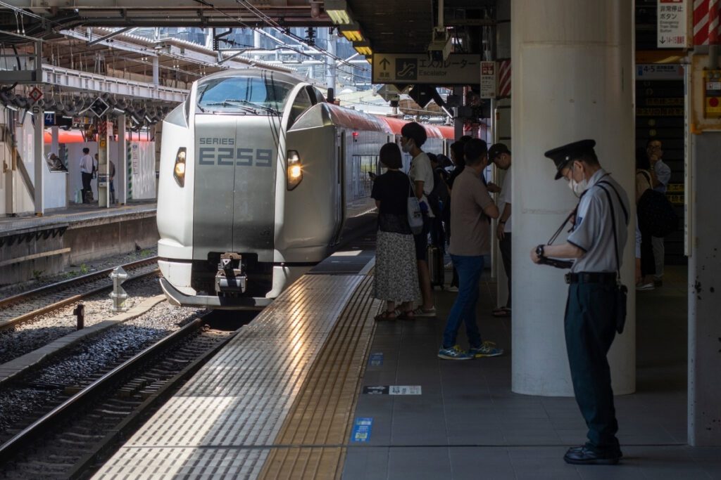 The Narita Express E259 series train arrives at Shinagawa Station, where passengers wait on the platform and a uniformed station staff member stands nearby checking his device.