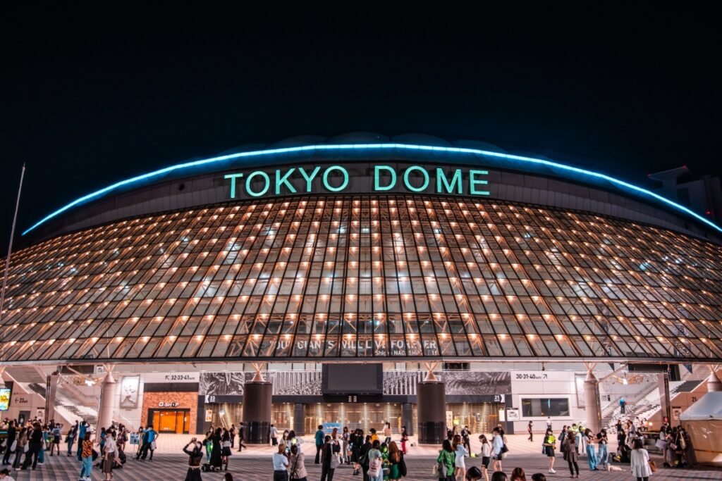 The image shows the brightly lit exterior of the Tokyo Dome at night, with its iconic curved glass facade and the words "TOKYO DOME" glowing in green neon at the top. A large crowd of people is gathered outside the entrance, creating a lively atmosphere typical of a major event or game at the stadium.