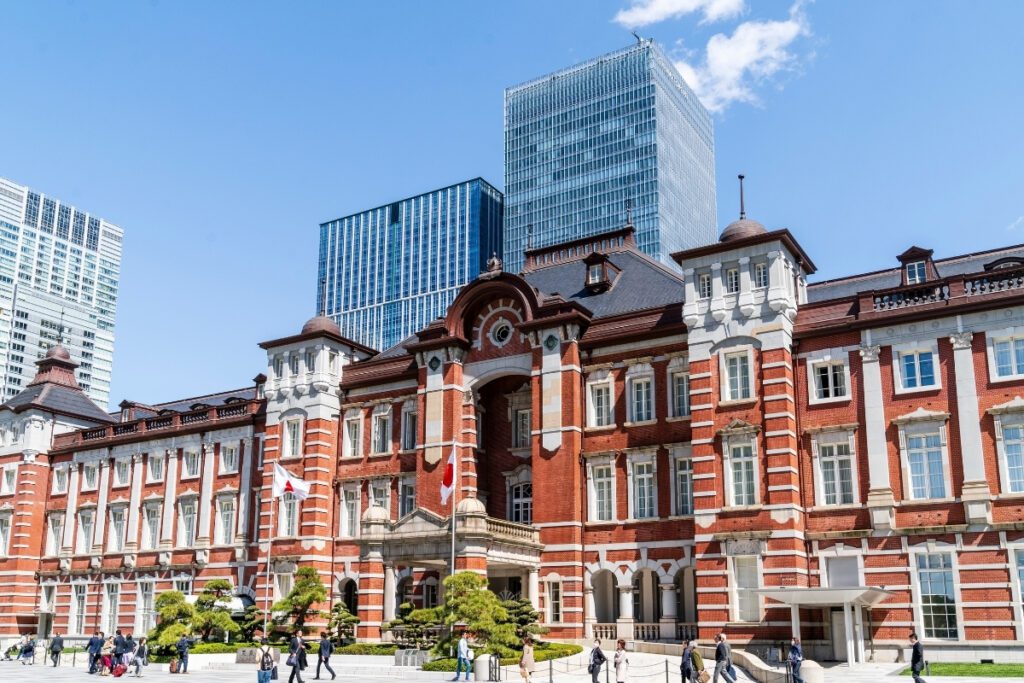 The historic red-brick facade of Tokyo Station stands in contrast to the modern skyscrapers behind it, with people walking in the foreground on a clear, sunny day.