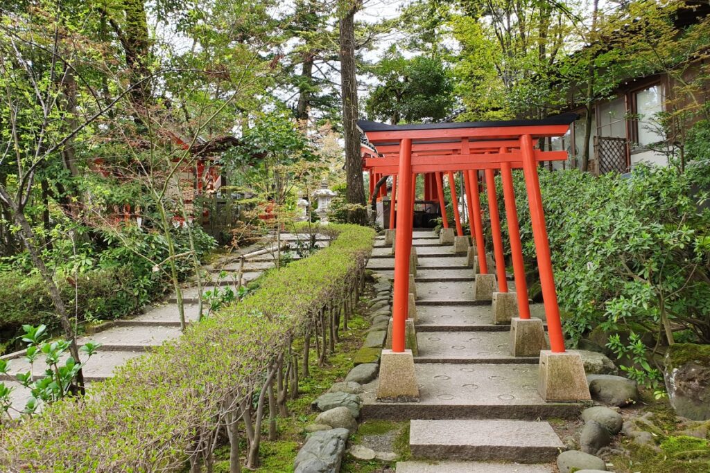 A quiet stone path at Kanazawa Shrine winds through lush greenery and passes beneath a series of small, vibrant red torii gates, creating a peaceful and atmospheric scene within a traditional Japanese garden.