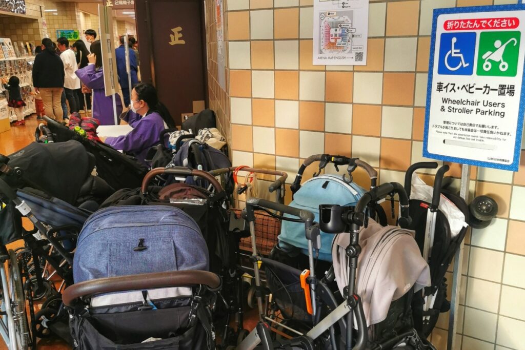 A designated wheelchair and stroller parking area inside Ryogoku Kokugikan arena, with a sign in Japanese and English indicating the space. Several strollers and mobility aids are parked against a tiled wall near a restroom entrance, while event staff in purple uniforms assist guests nearby.