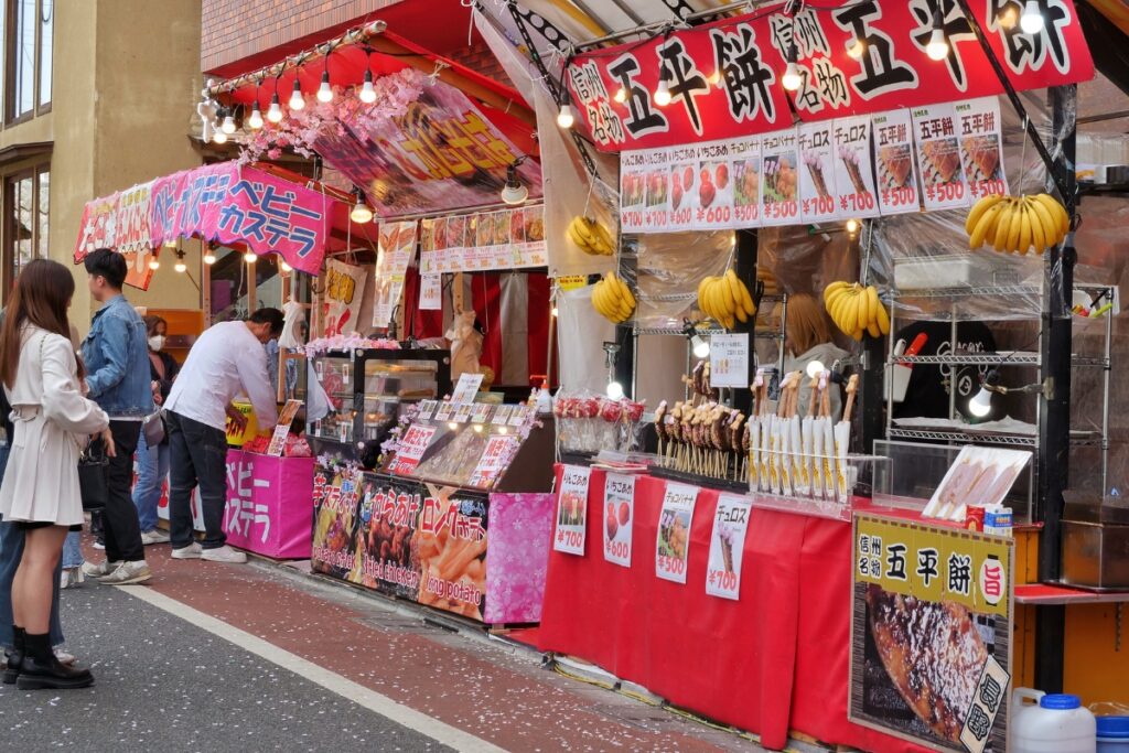 A colourful row of food stalls at Nakameguro during cherry blossom season, selling popular festival treats like candied apples, chocolate-covered bananas, mochi, fried chicken, and skewered snacks, with customers lining up along the petal-covered street.