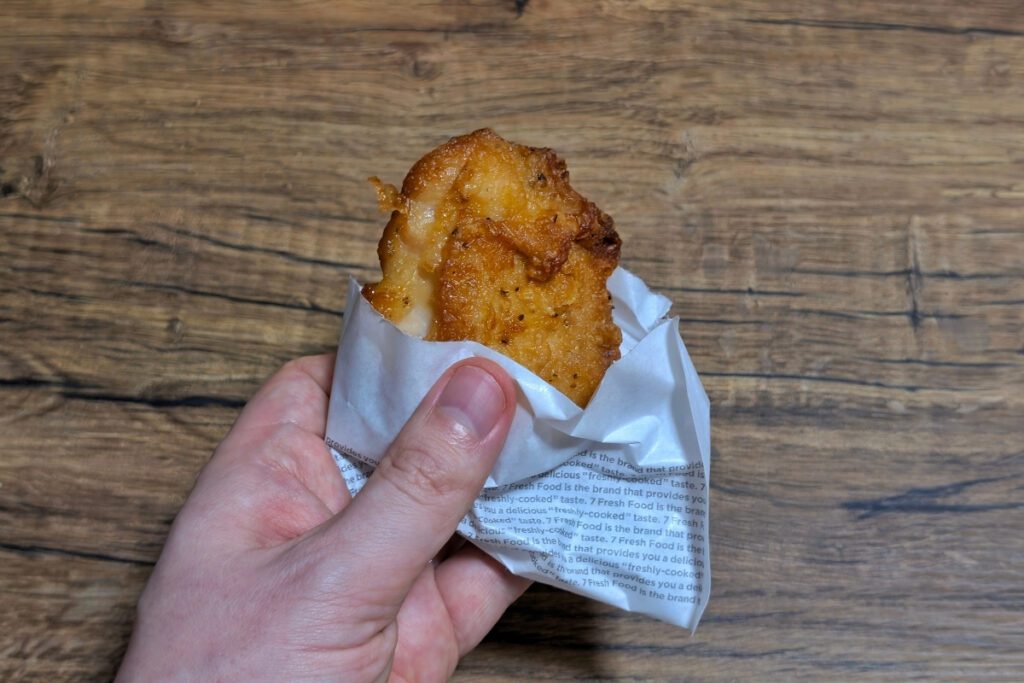 A hand holding a piece of golden-brown fried Nana Chiki chicken from Seven Eleven Japan wrapped in white paper with printed text, set against a wooden table background.