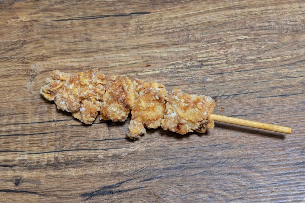 A wooden skewer with several pieces of golden-brown fried chicken, laid on a wooden surface. The crispy coating is textured with visible seasoning and crunchy bits.