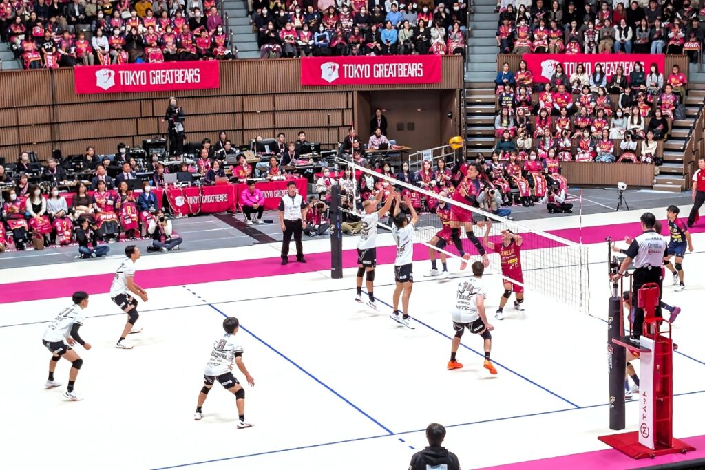 A Tokyo Great Bears player in a red jersey jumps high above the net to spike the ball, while three Nagano Tridents players in white jerseys leap to block. The packed arena, with fans in red and pink seated behind the court, watches closely.