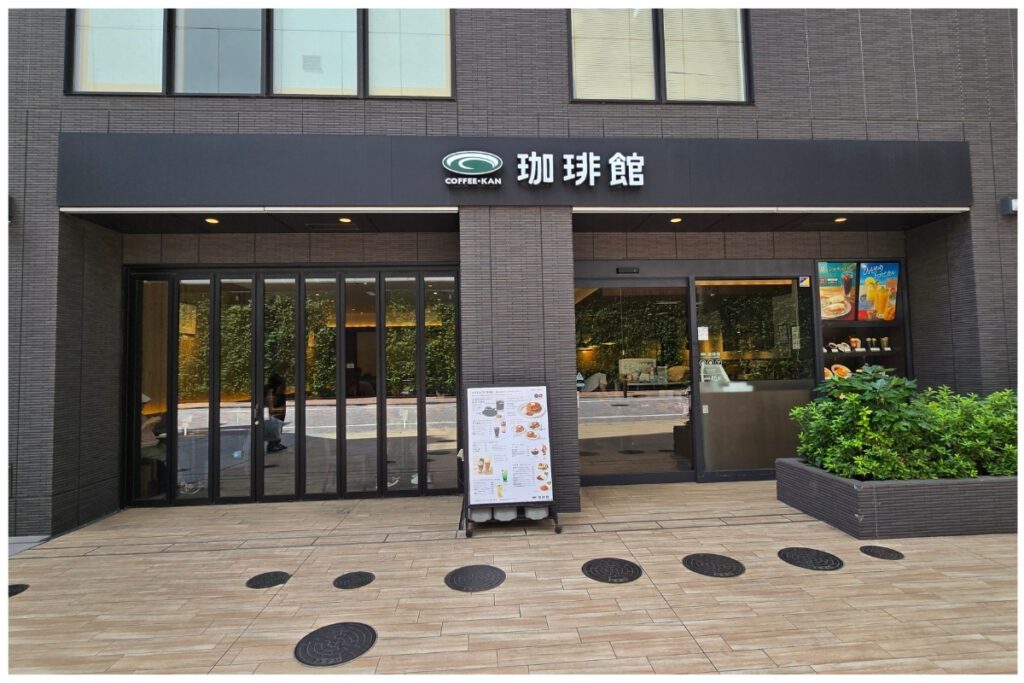 Coffee-Kan café on the ground floor beside Tosei Hotel Cocone Ueno Okachimachi, with glass frontage, a menu board outside, and indoor green plant wall visible through the windows.