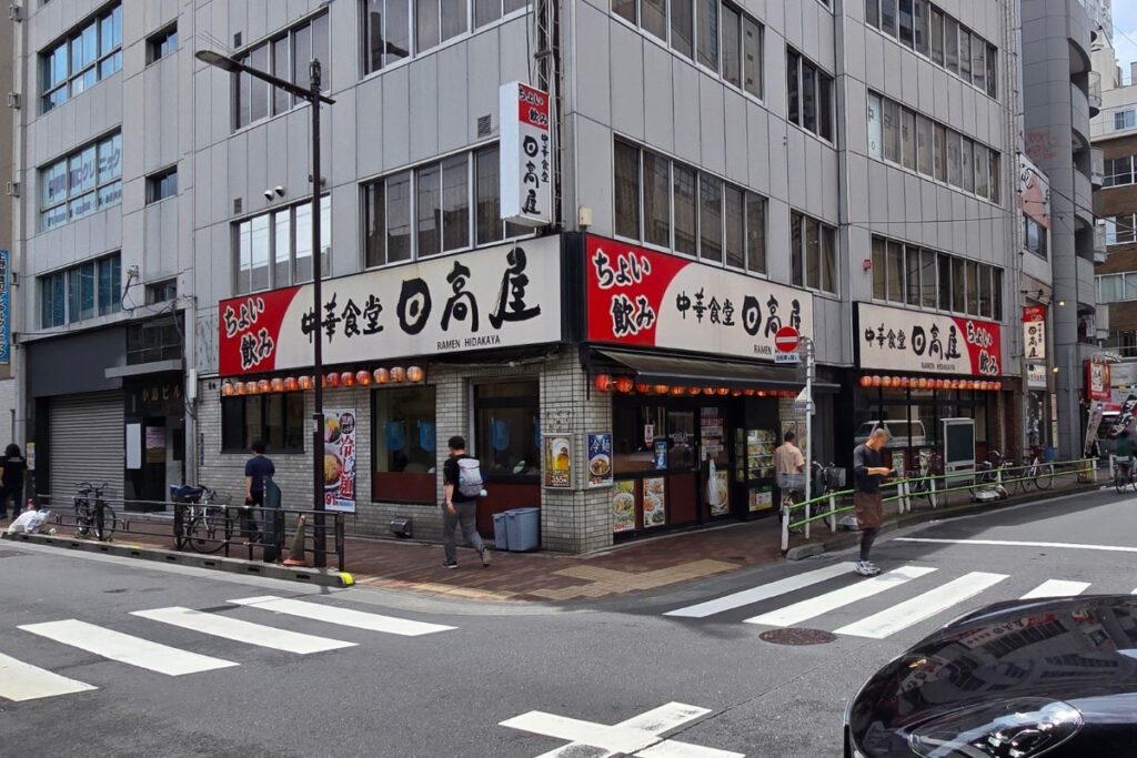 Ramen Hidakaya restaurant on a street corner directly beside Tosei Hotel Cocone Ueno Okachimachi, with red signage, paper lanterns, and pedestrians passing on the footpath.