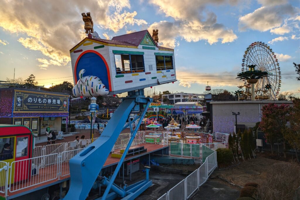 The Miracle Wan Room ride at Yomiuriland, a tilted house on a crane arm, with the Ferris wheel and park at sunset.