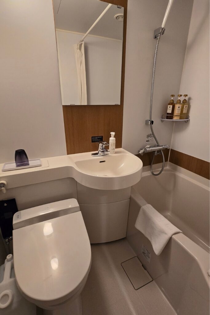 Compact modular bathroom at Dormy Inn Ueno Okachimachi with combined shower and bath, bidet toilet, wall-mounted sink, large mirror, timber wall panel, and toiletry bottles on a shower shelf.