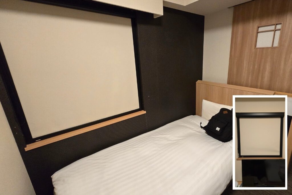 Fully closed blackout blind system in a single room at Dormy Inn Ueno Okachimachi, covering the entire wall beside the bed, with an inset showing the blind partially raised above the TV unit.