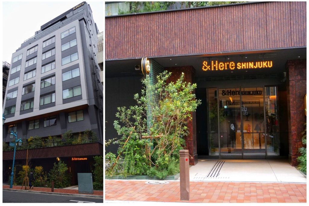 &Here Shinjuku hotel exterior showing a dark grey multi-storey facade with rust-coloured cladding, lush green plantings, and the illuminated &Here Shinjuku entrance sign above the glass doorway.