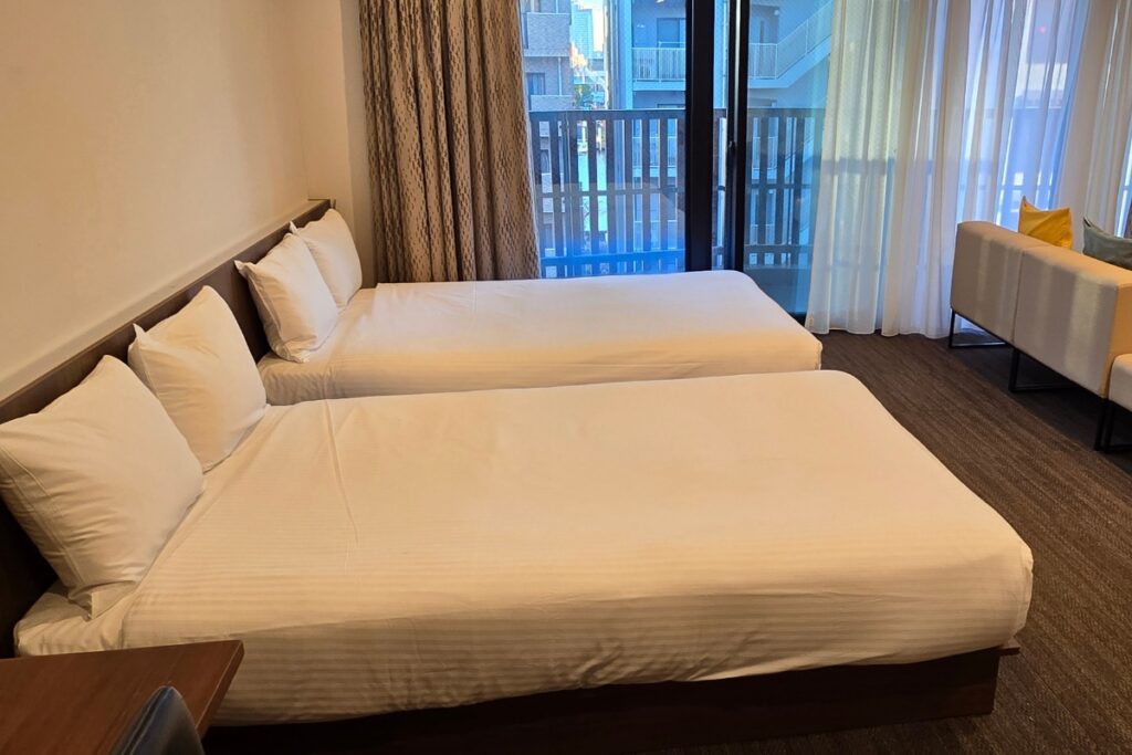 Alt text: Minn Asakusa apartment bedroom with two semi-double beds, floor-to-ceiling balcony doors with sheer curtains, and a sofa visible in the corner of the room.