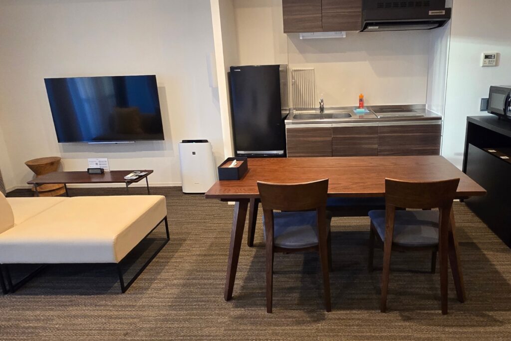 Minn Asakusa apartment living area with sofa, wall-mounted TV, dining table with two chairs, full-size kitchen with fridge and sink, microwave, and air purifier.