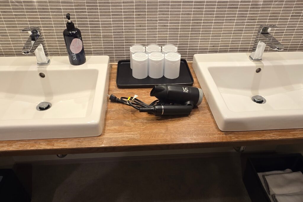 Minn Asakusa separate vanity area with two sinks on a timber countertop, hair dryer, toiletries, and mosaic tile splashback.